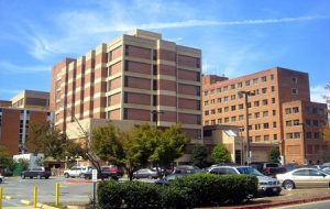 Large brick hospital complex with multiple buildings, a parking lot, and cars under a clear blue sky.