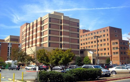 Large brick hospital complex with multiple buildings, a parking lot, and cars under a clear blue sky.