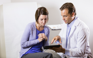 A doctor in a white coat discusses information on a tablet with a concerned female patient in an exam room.