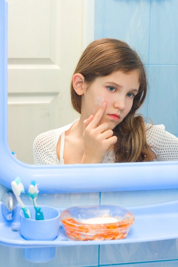 A young girl examining her face in the mirror, touching a blemish in a blue-themed bathroom.