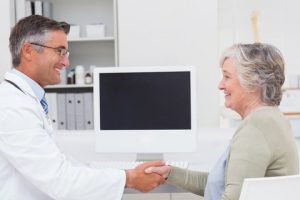 A doctor in a white coat shakes hands with an elderly woman in an office with a computer in the background.