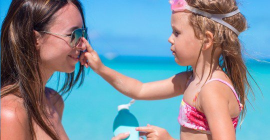 A young girl applying sunscreen to her mother's nose on a sunny beach.