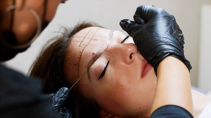 The process of shaping eyebrows by a beauty master with a thread for a young lady in the studio before the microblading. Close up view of a face while the procedure of marking lines.