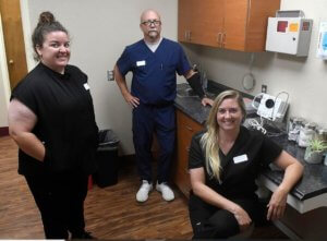 Three medical professionals in scrubs posing in a clinical setting with cabinets, a sink, and equipment.