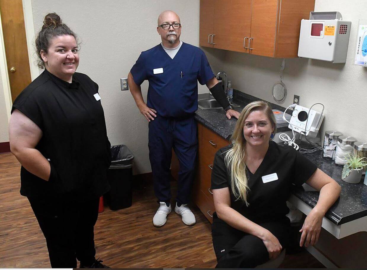 Three medical professionals in scrubs posing in a clinical setting with cabinets, a sink, and equipment.