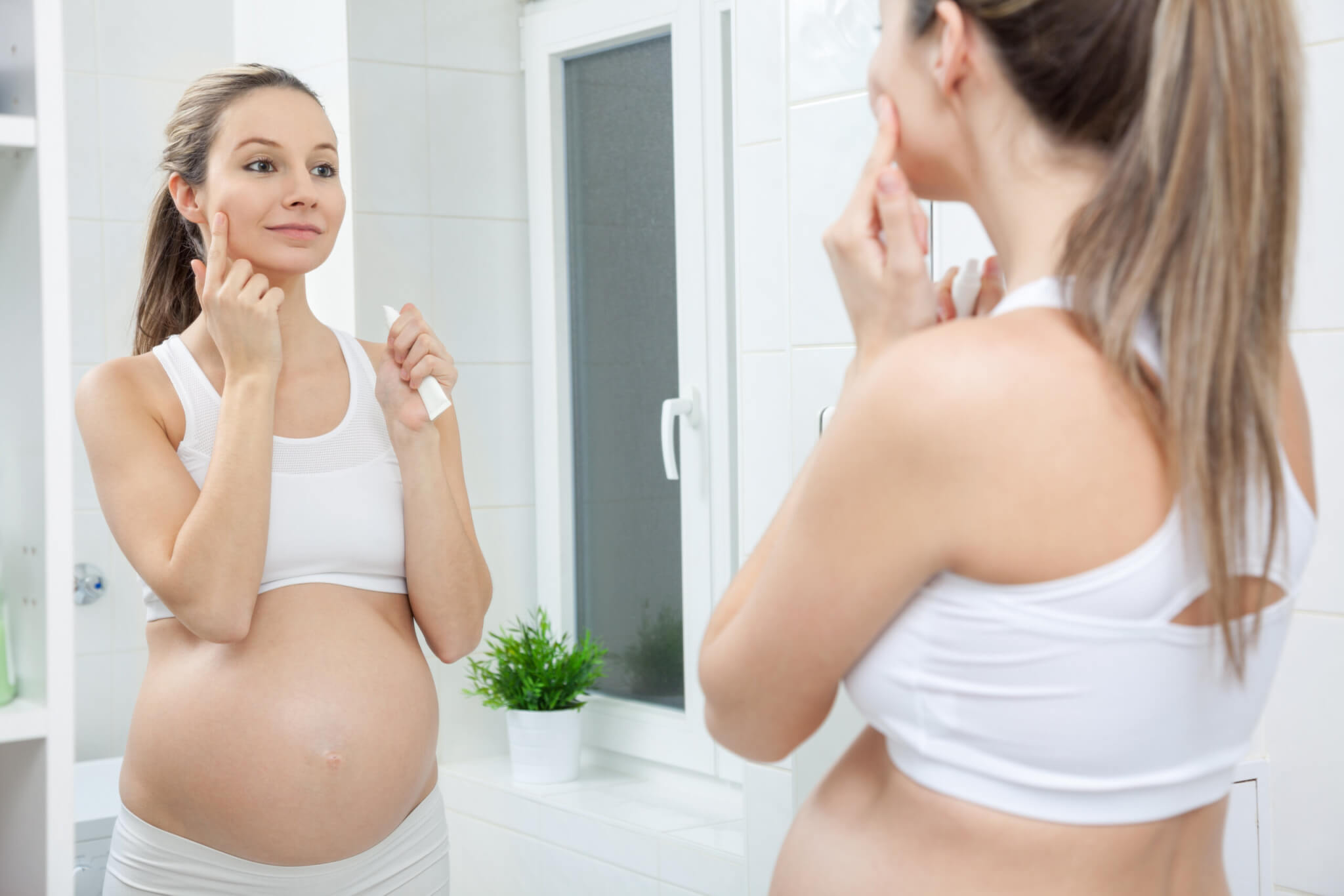 A pregnant women in a white sports bra applies skincare cream to her face while looking in the mirror.