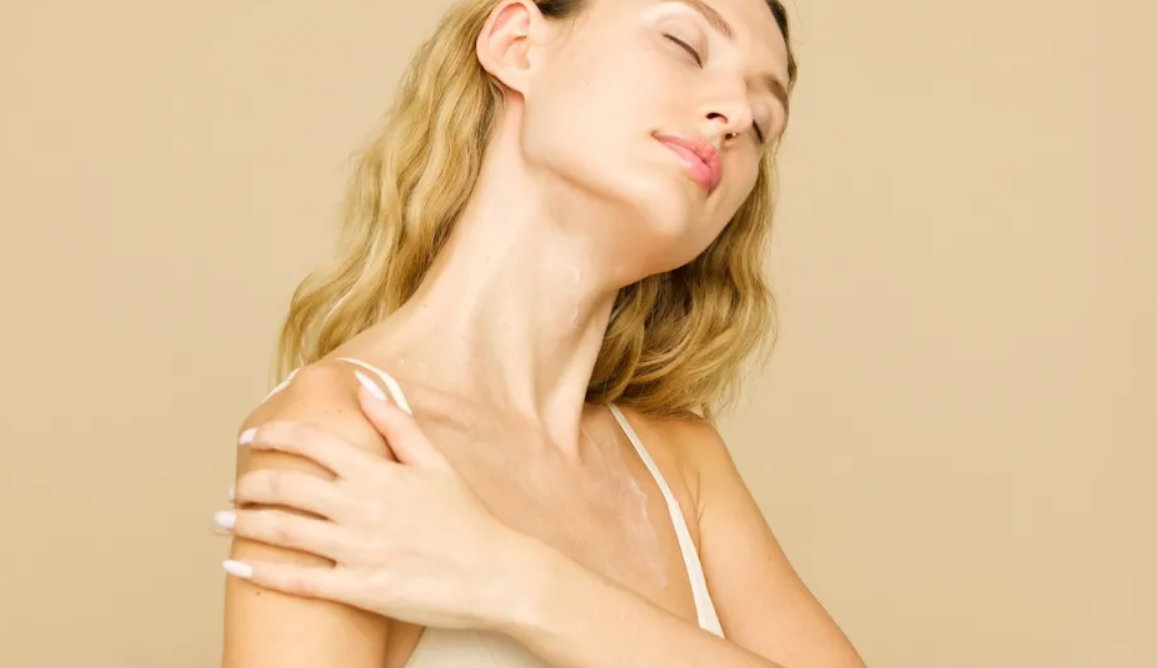 A woman with glowing skin applying lotion to her shoulder, enjoying a self-care moment.